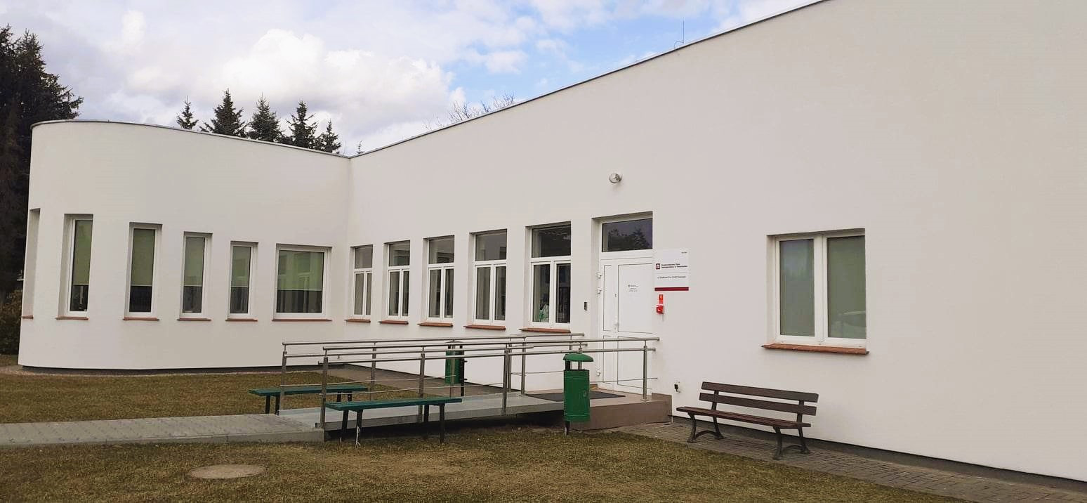 Nowy biały budynek, prosty w stylu nowoczesnym, z rampą do podjechania do wejścia. Przy ścianie i wejściu stoją kosze na śmieci i drewniane ławki