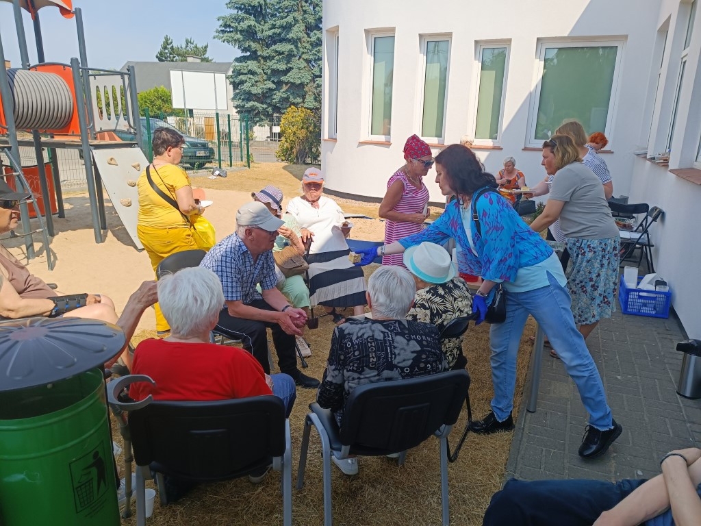 Grupa seniorów siedzi na krzesłach na zewnątrz, przy budynku z białymi ścianami i dużymi oknami. Część osób spożywa posiłki, inne rozmawiają. Wolontariusze i pracownicy w kolorowych ubraniach pomagają w obsłudze wydarzenia. W tle widać plac zabaw i ogrodzenie.