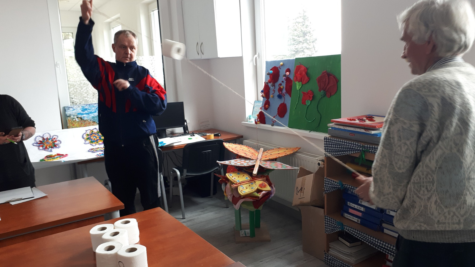 Zdjęcie przedstawia dwie osoby uczestniczące w grze lub zabawie, trzymające sznurek przepleciony przez rolkę papieru toaletowego. Na stole obok nich widoczne są kolejne rolki papieru. W pokoju znajdują się różne rękodzieła w kształcie kwiatów oraz motyli.