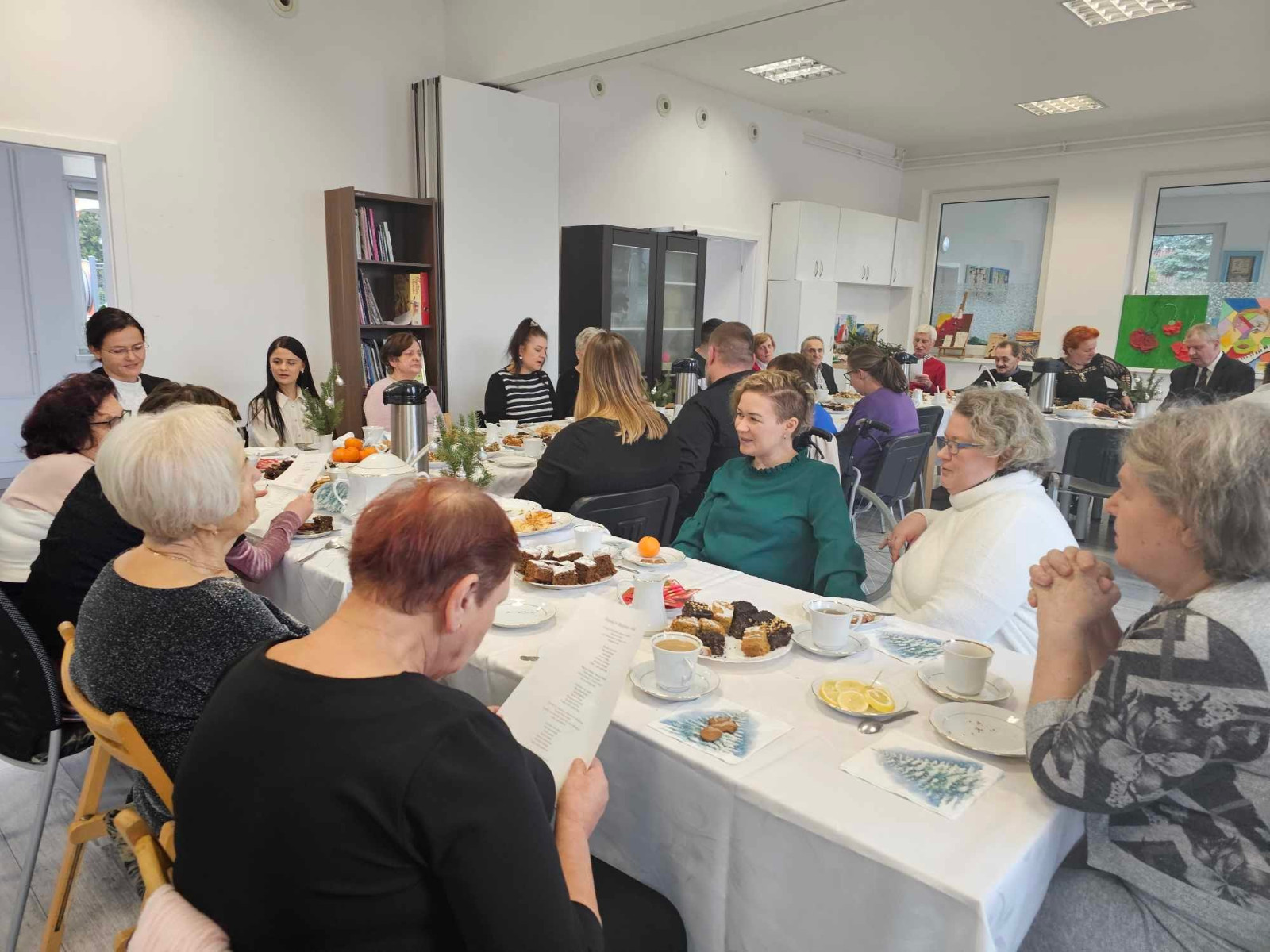 większa grupa ludzi, głównie seniorów, siedzi przy długim stole nakrytym białym obrusem. Na stole są ciasta, herbata, kawa, pomarańcze i dekoracje świąteczne. Część osób czyta kartki z tekstem, niektórzy prowadzą rozmowy.