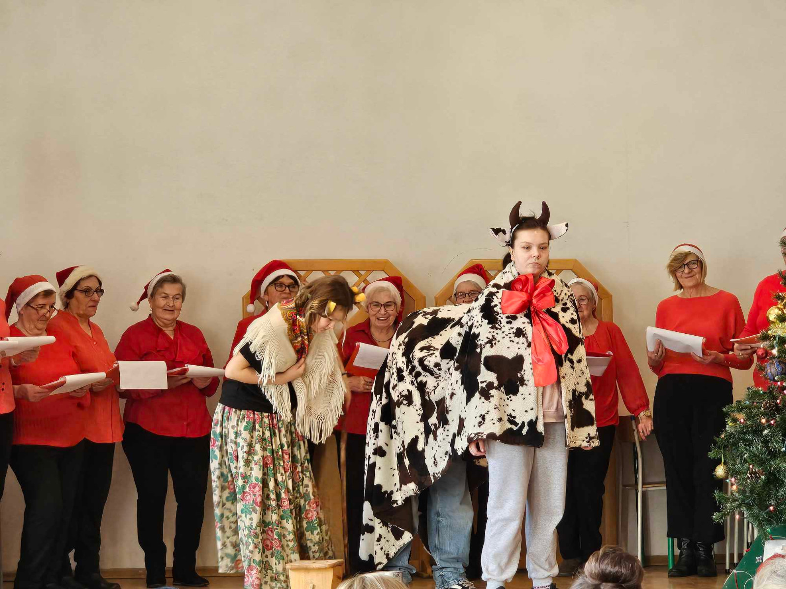 Kolędnicy na scenie a przed nimi scenka odgrywana, osoba przebrana za kobietę i dwie osoby za krowę