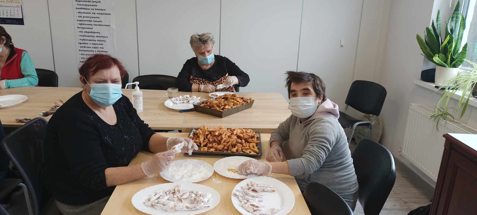 Trzy osoby w maseczkach i rękawiczkach przygotowują słodkie wypieki przy dużym stole. Na pierwszym planie kobieta w czarnej bluzce i mężczyzna w szarej bluzie obtaczają ciastka w cukrze pudrze, obok nich leżą talerze z gotowymi wypiekami. W tle starsza kobieta przygotowuje kolejne porcje na tacy.