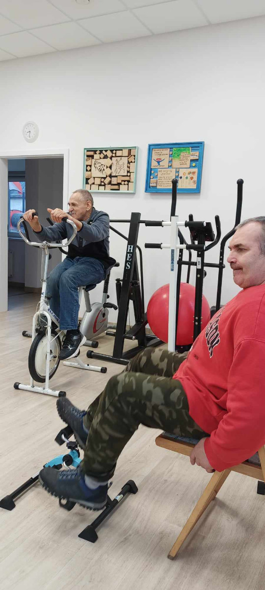 Dwóch mężczyzn korzysta ze sprzętu do ćwiczeń w sali rehabilitacyjnej. Jeden starszy mężczyzna w ciemnej kamizelce i jeansach pedałuje na rowerku stacjonarnym, a drugi, ubrany w czerwoną bluzę i spodnie moro, siedzi na krześle i korzysta z mini rowerka do ćwiczeń nóg. W tle sprzęty sportowe i dekoracje ścienne.