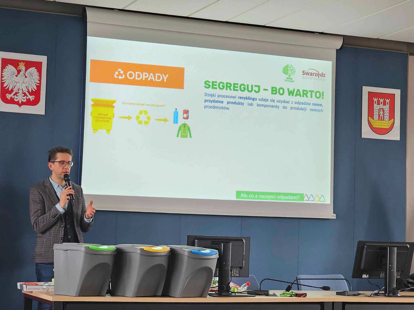 Mężczyzna w garniturze z mikrofonem stoi przed ekranem, na którym wyświetlana jest prezentacja na temat segregacji odpadów. Na stole znajdują się trzy pojemniki do segregacji śmieci. Na ekranie widoczny jest napis „Segreguj – bo warto!”.