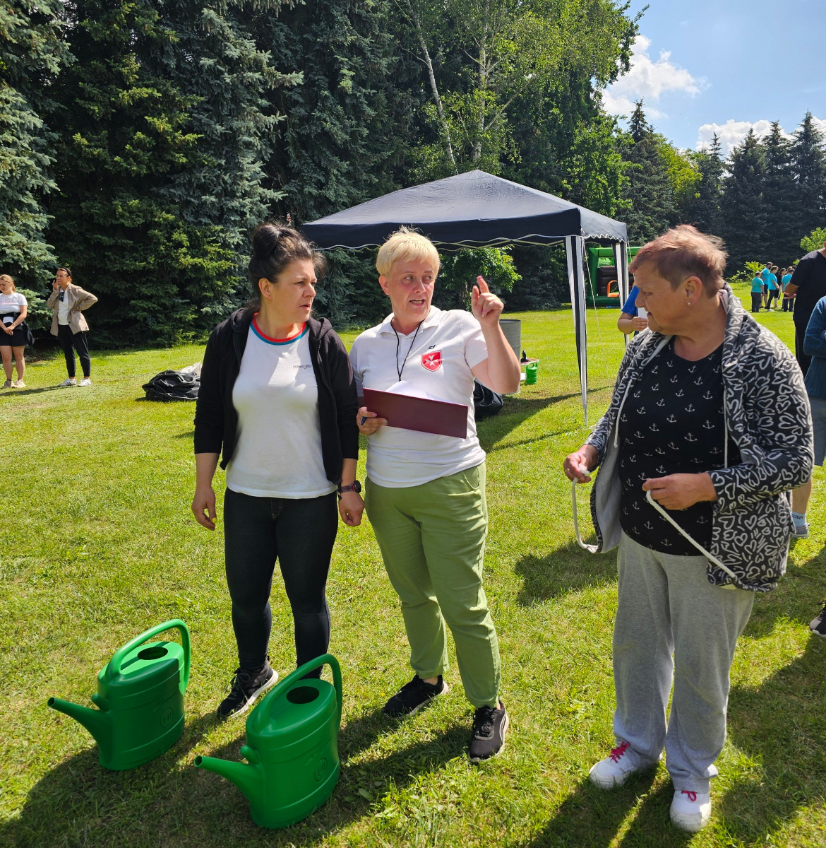 Trzy kobiety stoją na trawie, jedna z nich, sędzia, tłumaczy im zasady konkurencji sportowej, przed nimi stoją dwie zielone konewki które są elementem konkurencji. W tle widać namiot i innych ludzi uczestniczących w zawodach. Świeci słońce i jest ładna pogoda