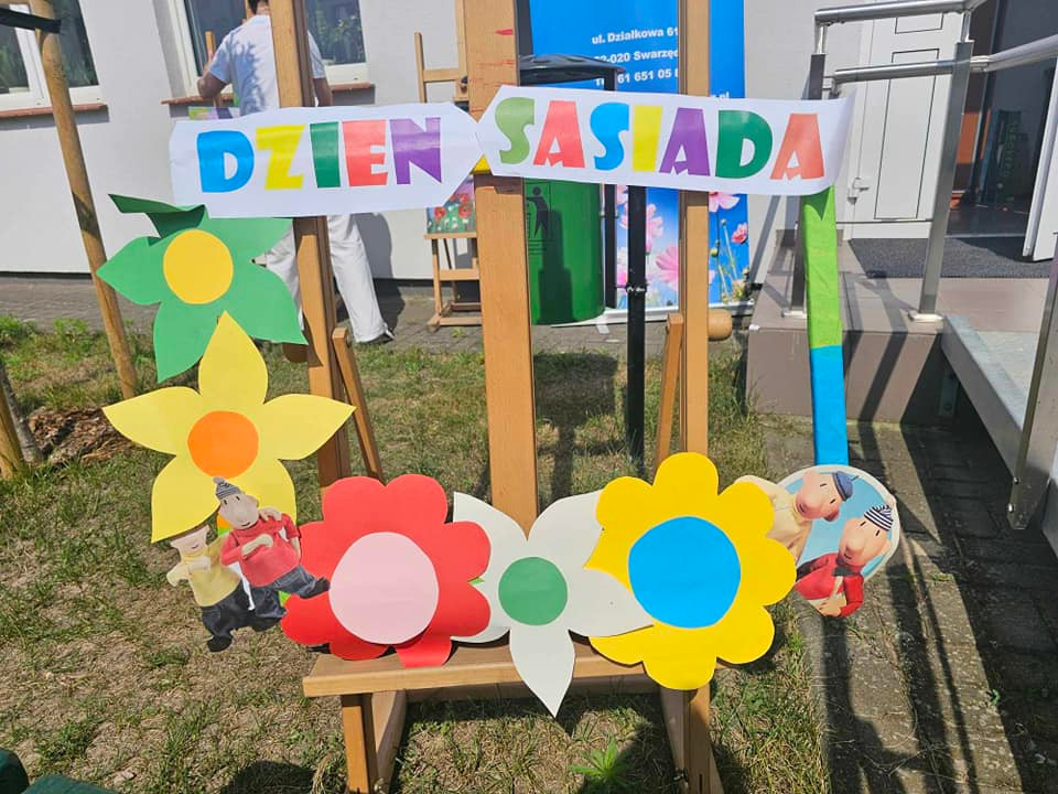 Kolorowa dekoracja z napisem „Dzień Sąsiada” umieszczona na drewnianym stelażu, ozdobiona papierowymi kwiatami i ilustracjami postaci. W tle fragment budynku oraz osoba w białym ubraniu