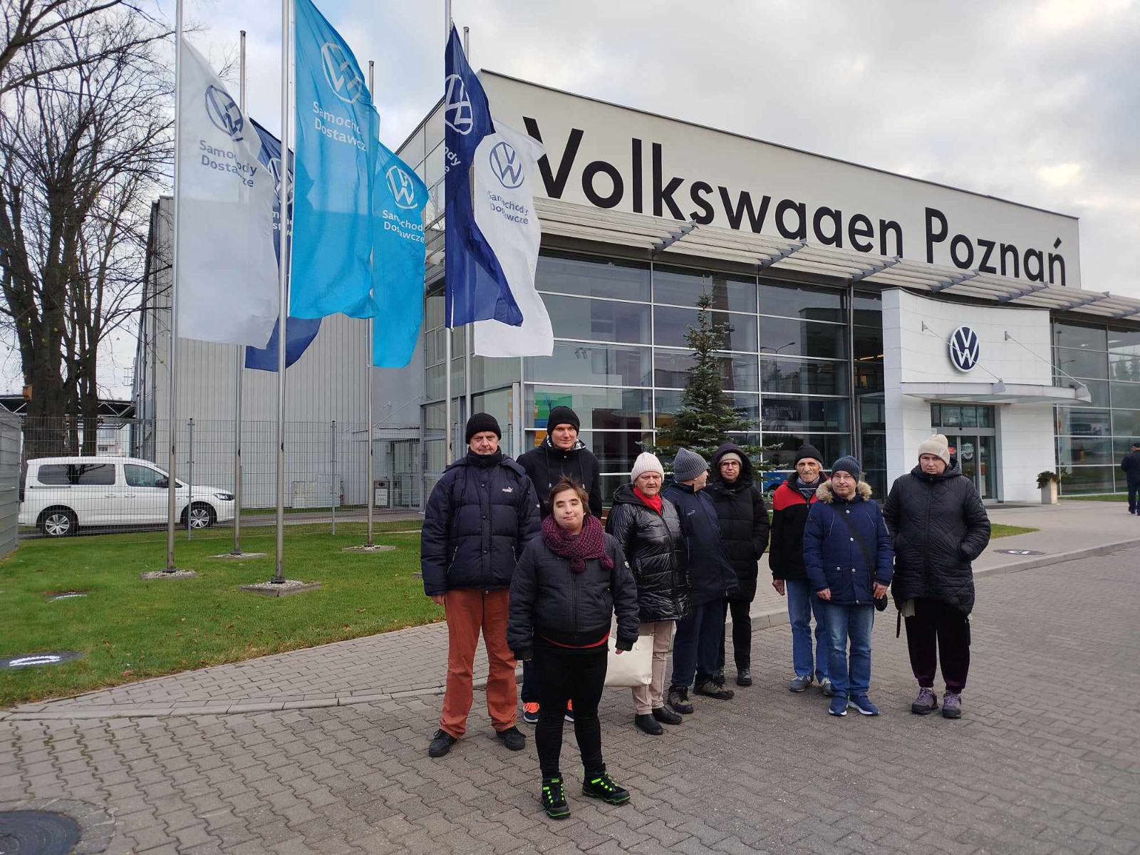 Grupa ciepło ubranych osób stoi przed wejściem do siedziby Volkswagen Poznań, za nimi powiewają flagi z logo firmy w różnych odcieniach niebieskiego i w kolorze białym. Za nimi widać trawnik i drogę dojazdową do firmy z białym samochodem
