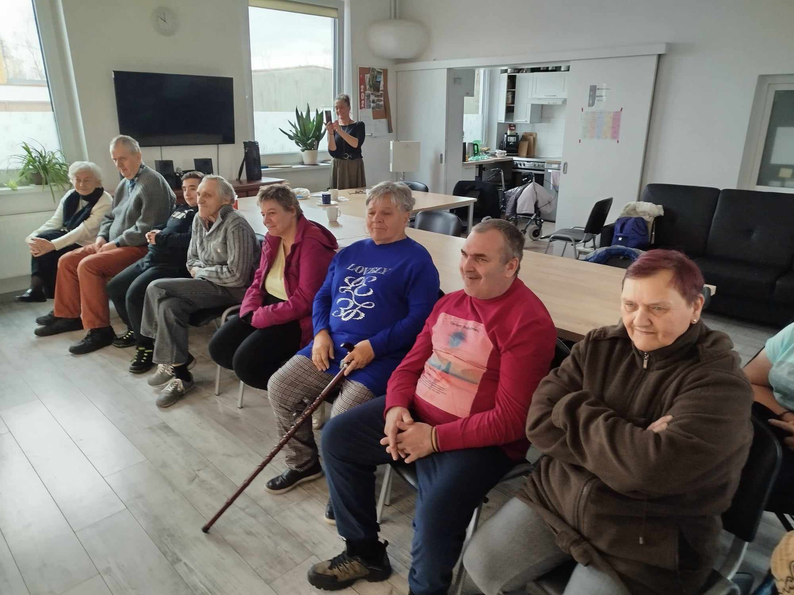 Grupa osób siedzących na krzesłach w sali, patrzących w jednym kierunku. Osoby uśmiechają się i z zaciekawieniem obserwują występ