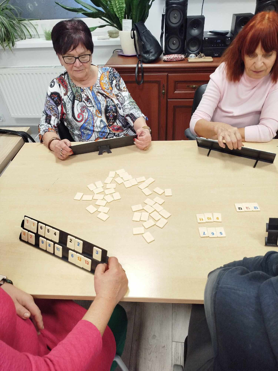 Cztery osoby siedzące przy stole podczas gry planszowej w Rummikub, z widocznymi płytkami na stojakach oraz na stole