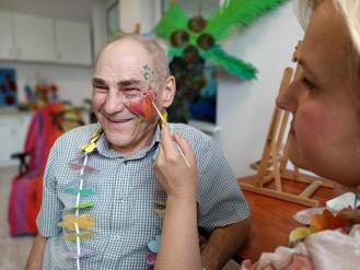 Starszy mężczyzna w koszuli w kratkę uśmiecha się, gdy kobieta maluje na jego twarzy kolorowy wzór kwiatowy. Ma na szyi dekoracyjny naszyjnik z wycinanych kwiatów. W tle widać kolorowe dekoracje i sztalugi, co sugeruje artystyczne lub integracyjne zajęcia.