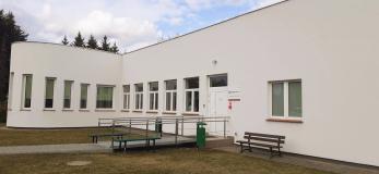 Nowy biały budynek, prosty w stylu nowoczesnym, z rampą do podjechania do wejścia. Przy ścianie i wejściu stoją kosze na śmieci i drewniane ławki 