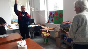 Zdjęcie przedstawia dwie osoby uczestniczące w grze lub zabawie, trzymające sznurek przepleciony przez rolkę papieru toaletowego. Na stole obok nich widoczne są kolejne rolki papieru. W pokoju znajdują się różne rękodzieła w kształcie kwiatów oraz motyli.