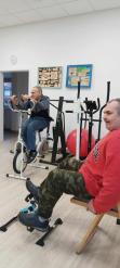 Dwóch mężczyzn korzysta ze sprzętu do ćwiczeń w sali rehabilitacyjnej. Jeden starszy mężczyzna w ciemnej kamizelce i jeansach pedałuje na rowerku stacjonarnym, a drugi, ubrany w czerwoną bluzę i spodnie moro, siedzi na krześle i korzysta z mini rowerka do ćwiczeń nóg. W tle sprzęty sportowe i dekoracje ścienne. 