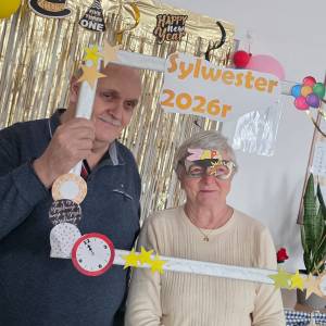 Mężczyzna i kobieta trzymają przed sobą białą drewnianą ramkę, udekorowaną imprezowo w papierowe gwiazdki, baloniki i brokat. Za nimi wisi na futrynie wisi złota brokatowa kurtyna.