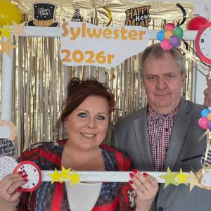 Mężczyzna i kobieta trzymają przed sobą białą drewnianą ramkę, udekorowaną imprezowo w papierowe gwiazdki, baloniki i brokat. Za nimi wisi na futrynie wisi złota brokatowa kurtyna.