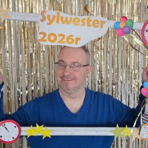 Mężczyzna trzyma przed sobą białą drewnianą ramkę, udekorowaną imprezowo w papierowe gwiazdki, baloniki i brokat. Za nim, na futrynie, wisi złota brokatowa kurtyna.