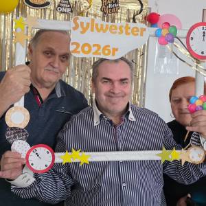 Dwoje mężczyzn i kobieta trzymają przed sobą białą drewnianą ramkę, udekorowaną imprezowo w papierowe gwiazdki, baloniki i brokat. Za nimi wisi na futrynie wisi złota brokatowa kurtyna.