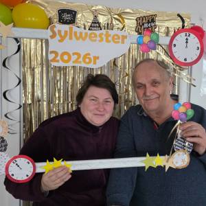 Mężczyzna i kobieta trzymają przed sobą białą drewnianą ramkę, udekorowaną imprezowo w papierowe gwiazdki, baloniki i brokat. Za nimi wisi na futrynie wisi złota brokatowa kurtyna.