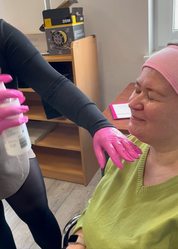 Uczestniczka ŚDS siedzi i mruży oczy podczas gdy kosmetolożka w różowych nitrylowych rękawiczkach spryskuje jej twarz za pomocą kosmetyku