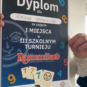Osoba trzyba w rękach dyplom chłopca za zajęcie pierwszego miejsca w trzecim szkolnym turnieju Rummikub