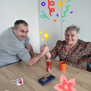 Mężczyzna i kobieta trzymają się dłońmi przy stole w pozycji jak gdyby mieli się siłować. Obydwoje są uśmiechnięci.