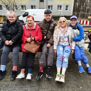 Pięć osób siedzących na ławce pozuje do zdjęcia z podwiniętymi nogawkami, pokazując kolorowe skarpetki