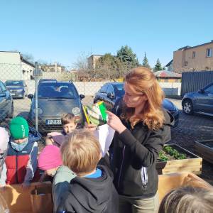 Prowadząca tłumaczy przedszkolakom proces sadzenia, trzymając w ręku saszetke z nasionkami