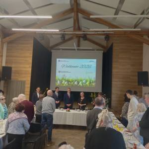 Zebranie osób w świetlicy. Na ścianie, za pomocą projektora, wyświetlana jest grafika Wielkanoc 2026 z logotypami organizujących spotkanie placówek. Za stołami z zastawą są uczestnicy ŚDS, Dzienniego Domu Pomocy Senior-Wigor oraz wybrane osoby z Gminy Swarzędz. 