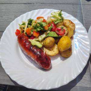 Zbliżenie na biały talerz z jedzeniem. Na talerzu widać przypieczoną na grillu kiełbasę, ziemniaczki oraz sałatkę z warzywkami