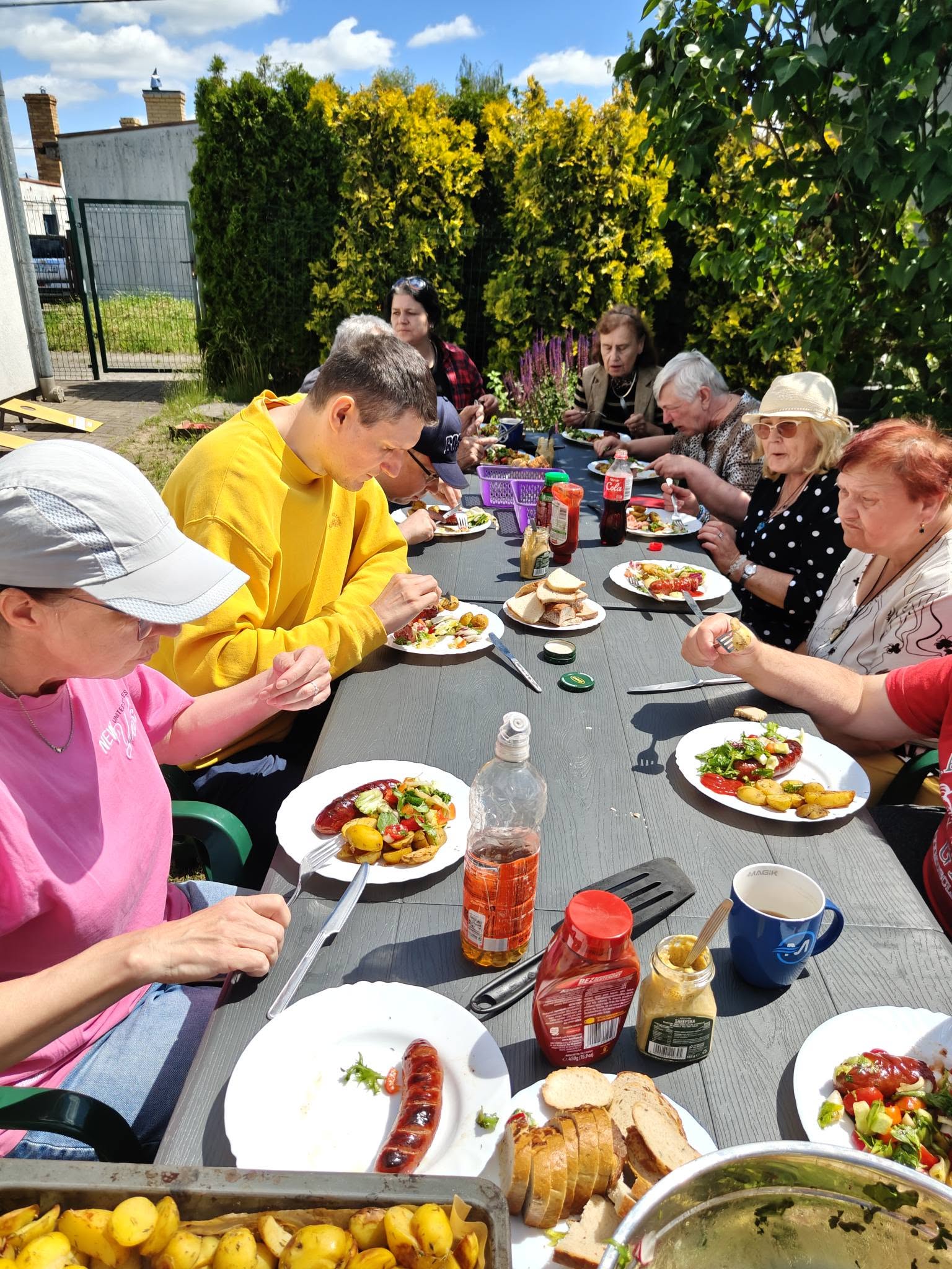 Uczestnicy ŚDS wspólnie świętują sezon grillowy przy długim stole. Stół wypełniony jest talerzami z jedzeniem i piciem