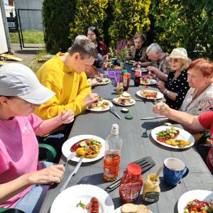 Uczestnicy ŚDS wspólnie świętują sezon grillowy przy długim stole. Stół wypełniony jest talerzami z jedzeniem i piciem
