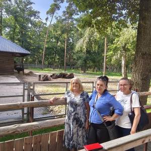 Trzy uśmiechnięte uczestniczki wycieczki pozują do pamiątkowego zdjęcia przy drewniano-metalowej barierce widokowej w pokazowej zagrodzie żubrów w Gołuchowie. W tle za ogrodzeniem widać stado żubrów odpoczywające na wybiegu przy drewnianej wiacie oraz wysokie leśne drzewa oświetlone promieniami słońca. Wspólny wyjazd integracyjny zorganizował Dzienny Dom „Senior-Wigor” w Swarzędzu we współpracy ze Środowiskowym Domem Samopomocy.
