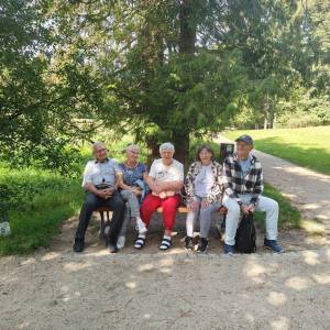 Grupa uśmiechniętych seniorów odpoczywa na drewnianej ławce parkowej w cieniu wysokiego drzewa iglastego podczas wycieczki do Gołuchowa. Pięcioro uczestników siedzi ramię w ramię przy żwirowej alejce, ciesząc się słonecznym dniem na łonie natury. W tle rozciąga się zielony, zadbany park krajobrazowy ze słoneczną polaną. Wyjazd integracyjny został zorganizowany przez Dzienny Dom „Senior-Wigor” w Swarzędzu we współpracy ze Środowiskowym Domem Samopomocy.   