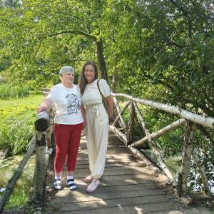 Dwie uśmiechnięte uczestniczki wycieczki pozują do zdjęcia na drewnianym mostku z brzozowymi poręczami, przerzuconym nad parkowym strumykiem w Gołuchowie. Kobiety stoją w otoczeniu bujnych, zielonych drzew i roślinności oświetlonych letnim słońcem. Wspólny integracyjny wyjazd rekreacyjny został zorganizowany przez Dzienny Dom „Senior-Wigor” w Swarzędzu oraz Środowiskowy Dom Samopomocy.