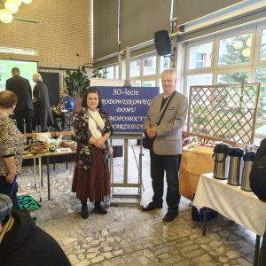 Kobieta w eleganckiej bordowej spódnicy i wzorzystym żakiecie oraz mężczyzna w szarej marynarce stoją obok niebieskiej tablicy na sztaludze z napisem „30-lecie Środowiskowego Domu Samopomocy w Swarzędzu”. Zdjęcie wykonano w jasnym holu Szkoły Podstawowej nr 3 podczas Światowego Dnia Zdrowia Psychicznego; w tle widać stoły z poczęstunkiem, tortem i termosami oraz uczestników wydarzenia.