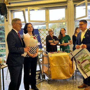 Burmistrz Swarzędza Marian Szkudlarek i II zastępca burmistrza Tomasz Zwoliński składają gratulacje kierownik Środowiskowego Domu Samopomocy Barbarze Chmieli z okazji 30-lecia placówki podczas Światowego Dnia Zdrowia Psychicznego w SP3 w Swarzędzu. Burmistrz wręcza bukiet kwiatów, zastępca trzyma pamiątkowy obraz z motywami Swarzędza, a w tle na wózku stoi udekorowany owocami tort jubileuszowy w obecności uśmiechniętych gości i pracowników.