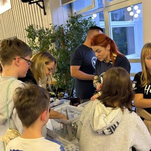 Dzieci i dorośli gromadzą się wokół stoiska warsztatowego Fundacji Błękitny Motyl podczas Światowego Dnia Zdrowia Psychicznego w Szkole Podstawowej nr 3 w Swarzędzu. Prowadzący prezentują uczestnikom przezroczyste pojemniki z materiałami edukacyjnymi, a obok widoczny jest baner z logo fundacji i duża roślina doniczkowa na tle okna.