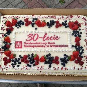 Duży, prostokątny tort jubileuszowy z okazji 30-lecia Środowiskowego Domu Samopomocy w Swarzędzu, umieszczony w kartonowym pudełku na wózku cateringowym. Wierzch pokryty jest białym kremem i ozdobiony czerwonym napisem okolicznościowym, logo placówki oraz obfitą dekoracją ze świeżych truskawek, malin i borówek amerykańskich wzdłuż brzegów. Tort przygotowano na obchody jubileuszu połączone ze Światowym Dniem Zdrowia Psychicznego w Szkole Podstawowej nr 3 w Swarzędzu.