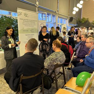 Prelegentka w ciemnym garniturze przemawia do grupy słuchaczy siedzących na krzesłach przed rollupem informacyjnym Centrum Zdrowia Psychicznego w trakcie Światowego Dnia Zdrowia Psychicznego w Szkole Podstawowej nr 3 w Swarzędzu. W jasnym holu widać stoiska konsultacyjne, materiały edukacyjne oraz innych uczestników wydarzenia rozmawiających w tle.
