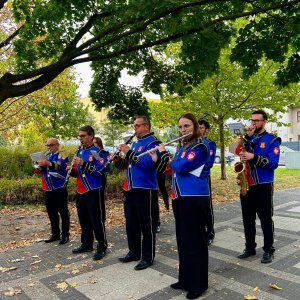 Muzycy Orkiestry Dętej ze Swarzędza w galowych, niebiesko-czerwonych mundurach z herbem miasta grają na instrumentach dętych (trąbka, klarnety, flet poprzeczny, saksofon) na wybrukowanej alejce pod zielonymi koronami drzew. Występ na świeżym powietrzu stanowił oprawę muzyczną obchodów Światowego Dnia Zdrowia Psychicznego oraz 30-lecia Środowiskowego Domu Samopomocy przy Szkole Podstawowej nr 3 w Swarzędzu.