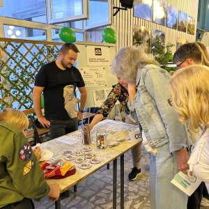 Uczestnicy wydarzenia, w tym harcerze w oliwkowych mundurach i dorośli, gromadzą się wokół stoiska warsztatowego Centrum Integracji Społecznej w Swarzędzu (CIS). Na stole leżą materiały plastyczne, słoiki, kredki oraz przygotowane do kolorowania podkładki z grafikami. W tle widnieje tablica informacyjna CIS z zielonymi balonami oraz duże okna holu Szkoły Podstawowej nr 3, w którym zorganizowano Światowy Dzień Zdrowia Psychicznego połączony z 30-leciem ŚDS.