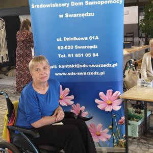Uśmiechnięta kobieta na wózku inwalidzkim, ubrana w niebieską bluzkę i ciemne spodnie, pozuje na tle dużego, niebieskiego rollupu informacyjnego Środowiskowego Domu Samopomocy w Swarzędzu. W tle widoczny jest hol Szkoły Podstawowej nr 3, stoiska warsztatowe oraz uczestnicy wydarzenia zorganizowanego z okazji Światowego Dnia Zdrowia Psychicznego połączonego z obchodami 30-lecia placówki.