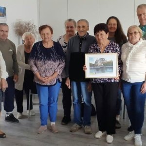 Pamiątkowe zdjęcie grupowe uczestników i opiekunów Środowiskowego Domu Samopomocy w Swarzędzu wraz z sąsiadką, panią Janiną, w jasnej sali placówki. Stojąca w centrum pani Janina trzyma oburącz oprawiony w jasną ramkę namalowany przez siebie obraz, przedstawiający ruiny Kopuły Bomby Atomowej w Hiroszimie nad brzegiem rzeki. Grupa uśmiecha się w stronę aparatu podczas spotkania podróżniczego połączonego z opowieściami o Japonii i wręczeniem podarowanego dzieła.