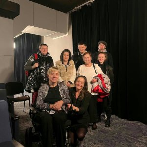 Pamiątkowe zdjęcie grupowe uczestników i opiekunów Środowiskowego Domu Samopomocy w Swarzędzu we wnętrzu Teatru Muzycznego w Poznaniu. Grupa pozuje uśmiechnięta na tle czarnych kotar teatralnych; na pierwszym planie siedzi kobieta na elektrycznym wózku inwalidzkim, obok której przykucnęła opiekunka, a za nimi stoją pozostali uczestnicy wyjścia. Zdjęcie wykonano podczas wyjazdu na musical „Deszczowa piosenka”, zorganizowanego w ramach współfinansowanego przez Samorząd Województwa Wielkopolskiego projektu „Teatr bez barier”.