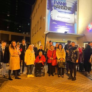 Pamiątkowe zdjęcie grupowe uczestników i opiekunów Środowiskowego Domu Samopomocy w Swarzędzu przed wejściem do Teatru Muzycznego po zmroku. Grupa w ciepłych, jesiennych płaszczach i kurtkach, w tym uśmiechnięta kobieta na wózku inwalidzkim w czerwonej kurtce na pierwszym planie, pozuje na wybrukowanym placu pod rozświetlonym, dużym plakatem spektaklu teatralnego. Wyjście na wydarzenie teatralne zorganizowano w ramach projektu „Teatr bez barier”, współfinansowanego ze środków Samorządu Województwa Wielkopolskiego.