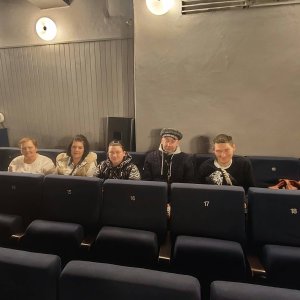 Pięcioro uczestników Środowiskowego Domu Samopomocy w Swarzędzu siedzi w rzędzie granatowych foteli na widowni Teatru Muzycznego, czekając na rozpoczęcie spektaklu i uśmiechając się w stronę aparatu. W tle widoczna jest szara ściana sali oświetlona okrągłymi lampami ściennymi. Wyjście na musical „Deszczowa piosenka” odbyło się w ramach projektu „Teatr bez barier”, współfinansowanego ze środków Samorządu Województwa Wielkopolskiego.