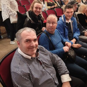 Uczestnicy i opiekunka ze Środowiskowego Domu Samopomocy w Swarzędzu siedzą na bordowych fotelach na widowni teatru, uśmiechając się w stronę aparatu przed rozpoczęciem spektaklu. Na pierwszym planie widoczny jest uśmiechnięty mężczyzna w pasiastej koszuli, obok niego siedzą dwaj koledzy, a z rzędu za nimi spogląda uśmiechnięta kobieta w czarnej kurtce i szalu. W tle widać zapełniające się rzędy widowni teatralnej. Zdjęcie wykonano podczas wyjścia na spektakl baletowy „Conrad” w ramach projektu „Teatr bez barier”, współfinansowanego ze środków Samorządu Województwa Wielkopolskiego.
