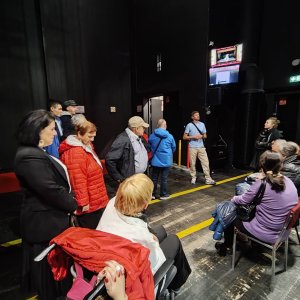 Grupa uczestników i opiekunów Środowiskowego Domu Samopomocy w Swarzędzu podczas zwiedzania kulis teatru z przewodnikiem. Pracownik instytucji w błękitnej koszulce stoi na deskach sceny i opowiada zebranym o kulisach spektaklu baletowego. Uczestnicy – część stojąca, część siedząca na krzesłach oraz kobieta na wózku inwalidzkim – słuchają prelekcji w przestrzeni technicznej pod zawieszonym ekranem monitoringu sceny. Wyjście na spektakl „Conrad” zorganizowano w ramach projektu „Teatr bez barier”, współfinansowanego ze środków Samorządu Województwa Wielkopolskiego.