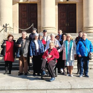 Pamiątkowe zdjęcie grupowe uczestników i opiekunów Środowiskowego Domu Samopomocy w Swarzędzu na schodach przed wejściem do Teatru Wielkiego im. Stanisława Moniuszki w Poznaniu, w pełnym słońcu. W centralnej części kadru siedzi uśmiechnięta kobieta na wózku inwalidzkim w czerwonej kurtce, otoczona przez grupę stojącą na kamiennych stopniach. W tle widoczne są masywne kolumny neoklasycystycznego portyku, drewniane wrota teatru oraz fragment rzeźby lwa. Zdjęcie wykonano podczas wyjścia na spektakl baletowy „Conrad” zrealizowanego w ramach projektu „Teatr bez barier”, współfinansowanego ze środków Samorządu Województwa Wielkopolskiego.
