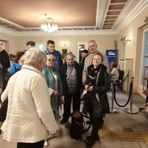 Grupa uczestników i opiekunów ze Środowiskowego Domu Samopomocy w Swarzędzu stoi w jasnym, eleganckim foyer teatru pod ozdobnym żyrandolem. Uczestnicy w wierzchnich okryciach rozmawiają ze sobą w oczekiwaniu na wejście na widownię, a po prawej stronie widoczne są stojaki z materiałami informacyjnymi i repertuarem. Zdjęcie dokumentuje wyjście na spektakl baletowy „Conrad” zorganizowane w ramach projektu „Teatr bez barier”, współfinansowanego ze środków Samorządu Województwa Wielkopolskiego.